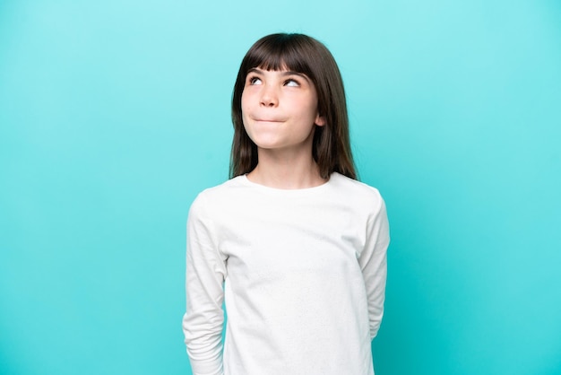 Little caucasian girl isolated on blue background having doubts while looking up