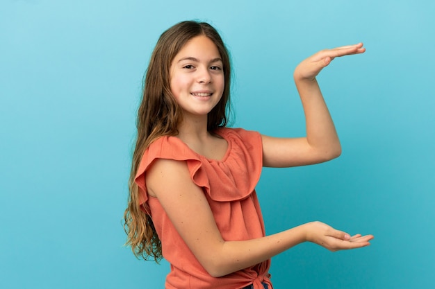 Little caucasian girl isolated on blue background holding copyspace to insert an ad