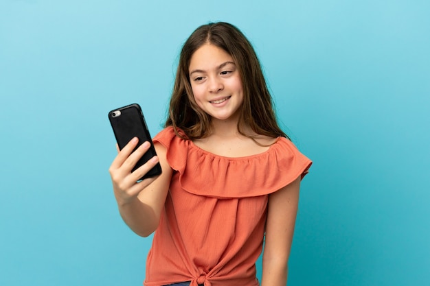 Piccola ragazza caucasica isolata su sfondo blu che fa un selfie