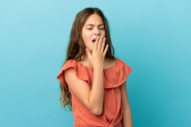 Little caucasian girl isolated on blue background yawning and covering wide open mouth with hand