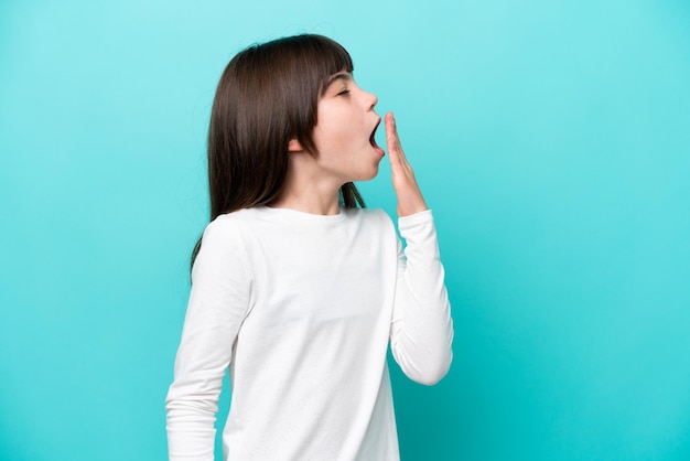 Little caucasian girl isolated on blue background yawning and covering wide open mouth with hand