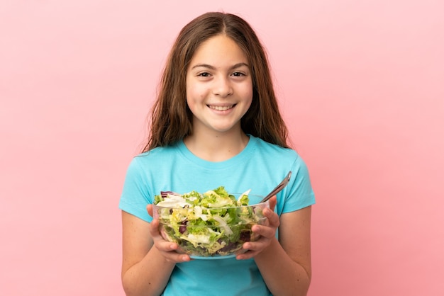 Little caucasian girl isolated on pink background holding a bowl of salad with happy expression