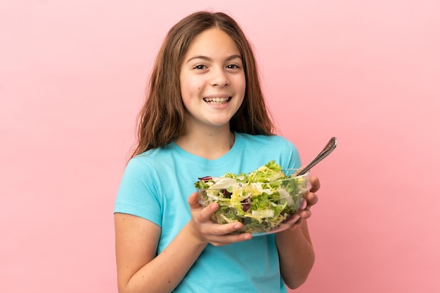 Little caucasian girl isolated on pink background holding a bowl of salad with happy expression