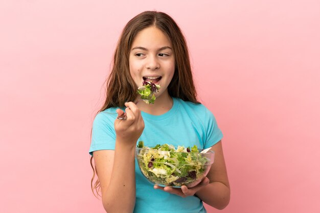 Little caucasian girl isolated on pink background holding a bowl of salad with happy expression