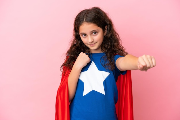 Little caucasian girl isolated on pink background in superhero costume and fighting