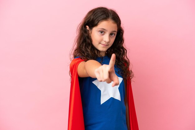 Little caucasian girl isolated on pink background in superhero costume and pointing to the front