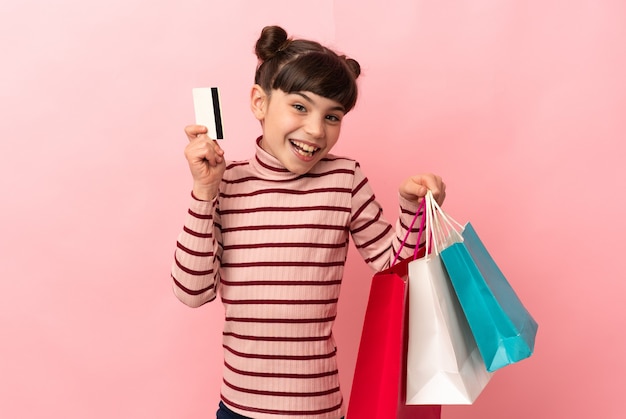 Piccola ragazza caucasica isolata sulla parete rosa che tiene i sacchetti della spesa e una carta di credito