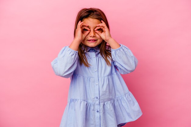 Little caucasian girl isolated on pink wall  showing okay sign over eyes