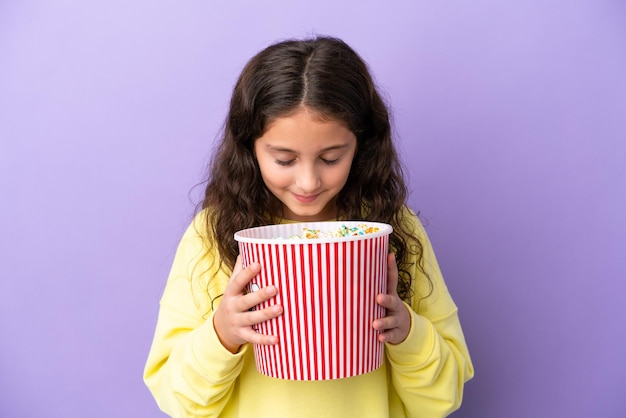 Little caucasian girl isolated on purple background holding a big bucket of popcorns