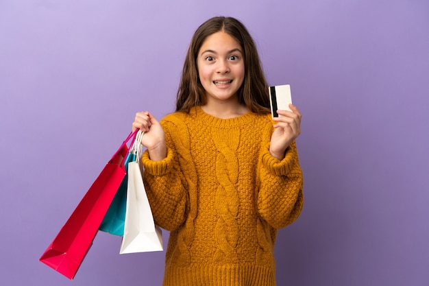 Little caucasian girl isolated on purple background holding shopping bags and surprised