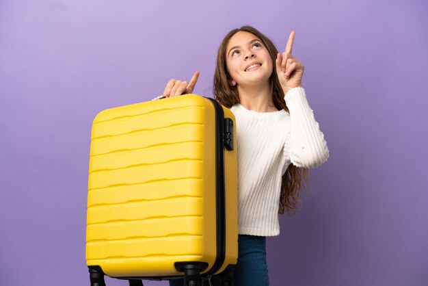 Photo little caucasian girl isolated on purple background in vacation with travel suitcase and pointing up