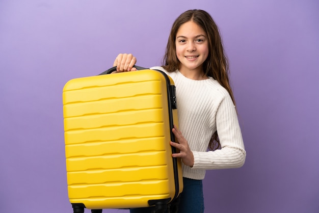 Little caucasian girl isolated on purple background in vacation with travel suitcase