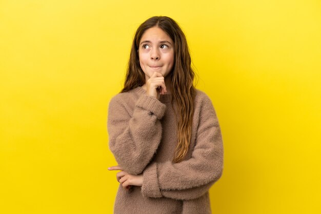 Little caucasian girl isolated on yellow background having doubts and thinking