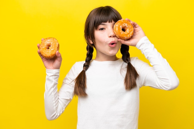 Little caucasian girl isolated on yellow background holding a donut in an eye