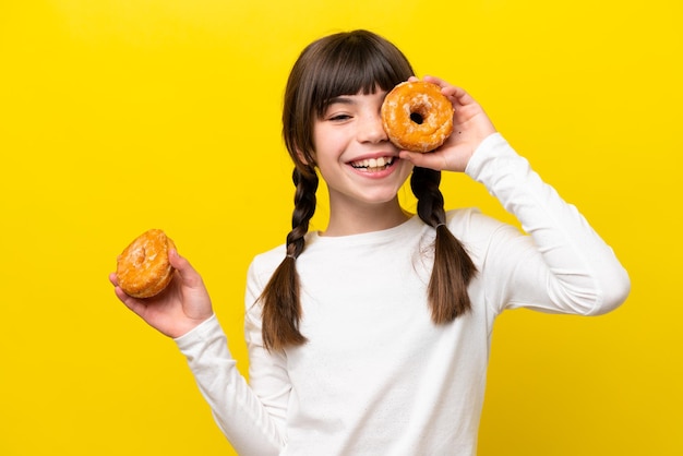 Little caucasian girl isolated on yellow background holding a donut and happy