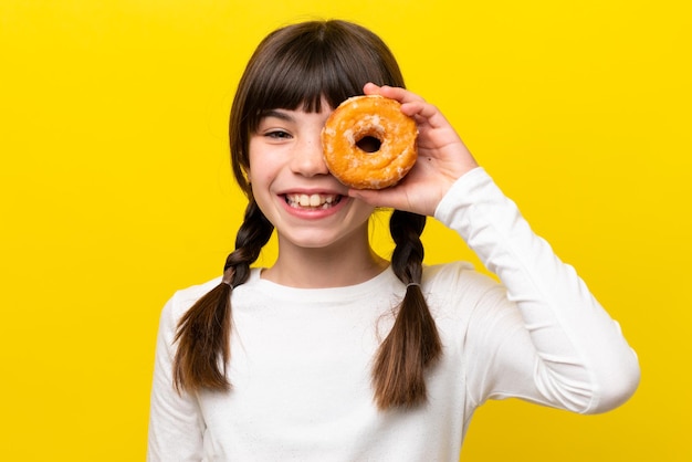 Little caucasian girl isolated on yellow background holding a donut and happy