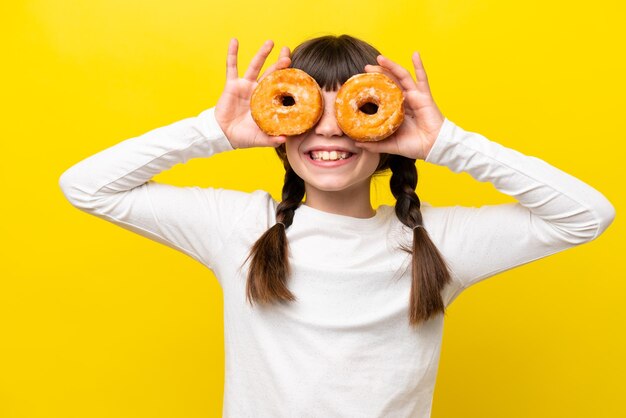 Little caucasian girl isolated on yellow background holding donuts in eyes