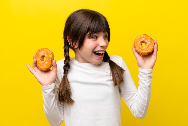 Little caucasian girl isolated on yellow background holding donuts and surprised