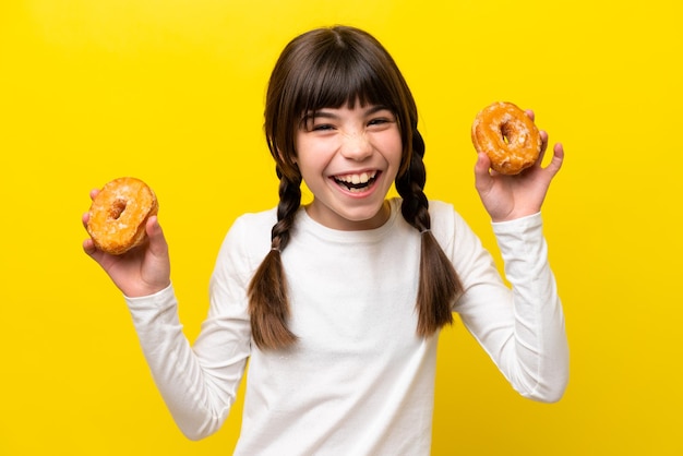 Little caucasian girl isolated on yellow background holding donuts with happy expression