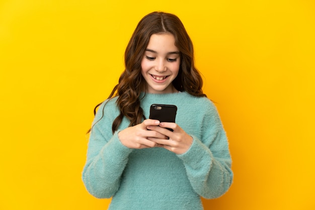 Little caucasian girl isolated on yellow background sending a message or email with the mobile