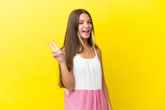 Little caucasian girl isolated on yellow background smiling and showing victory sign