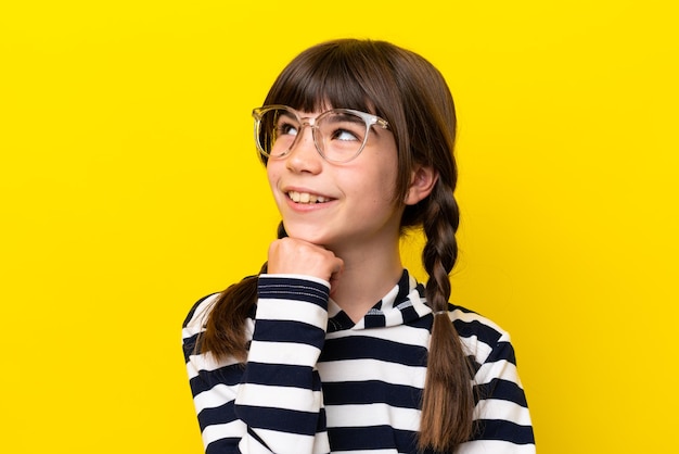 Little caucasian girl isolated on yellow background With glasses and thinking while looking up