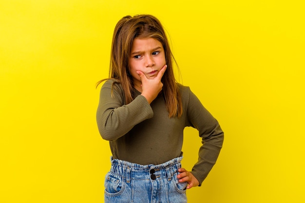 Little caucasian girl isolated on yellow contemplating, planning a strategy, thinking about the way of a business.