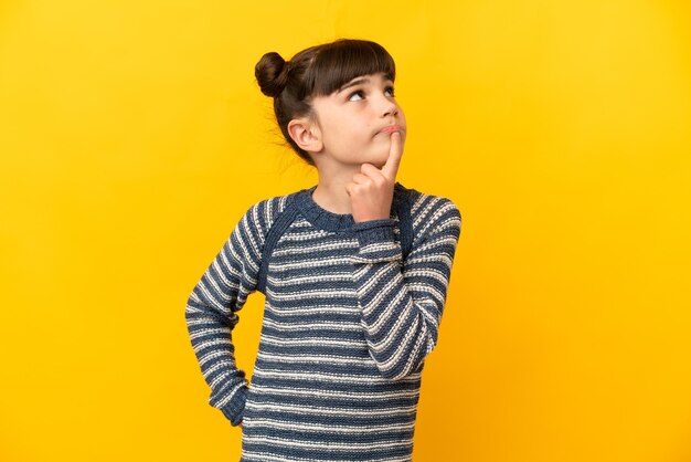 Little caucasian girl isolated on yellow having doubts while looking up