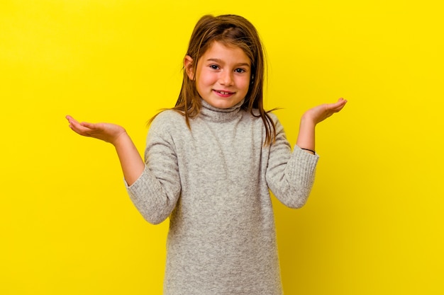 Little caucasian girl isolated on yellow wall makes scale with arms, feels happy and confident.