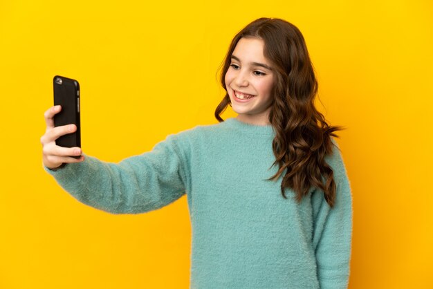 Little caucasian girl isolated on yellow wall making a selfie with mobile phone