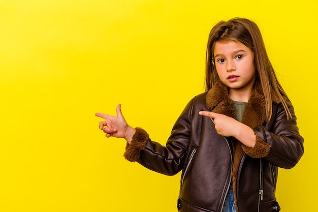 Little caucasian girl isolated on yellow wall shocked pointing with index fingers to a copy space.