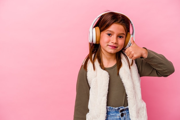 Little caucasian girl listening music isolated on pink background smiling and raising thumb up