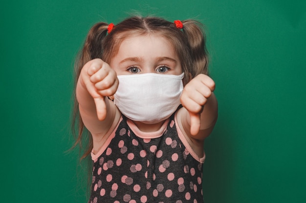 Little caucasian girl in medical mask and polka dot dress showing thumb sign on green background during quarantine and coronavirus pandemic 2020