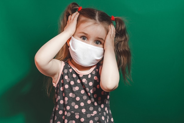 Little caucasian girl in medical mask wears red polka dot dress in studio on green background and holds her head in pain 2021