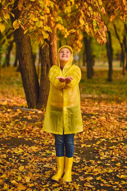 小さな白人の女の子は秋の雨の中で遊ぶ。屋外で自然で遊んでいる子供。女の子は黄色いレインコートを着て、雨を楽しんでいます。