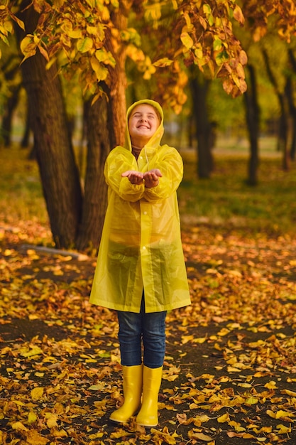 小さな白人の女の子は秋の雨の中で遊ぶ。屋外で自然で遊んでいる子供。女の子は黄色いレインコートを着て、雨を楽しんでいます。