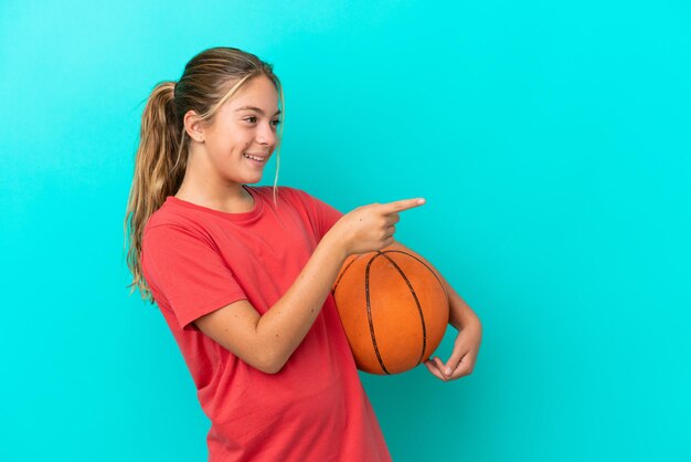 Little caucasian girl playing basketball isolated on blue background pointing finger to the side and presenting a product