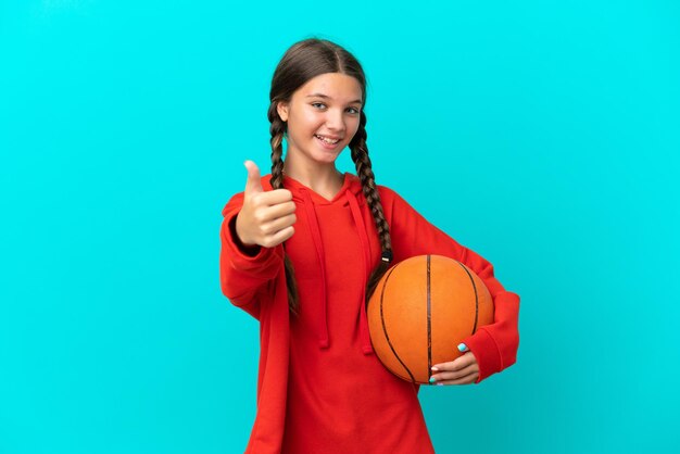 Little caucasian girl playing basketball isolated on blue background with thumbs up because something good has happened