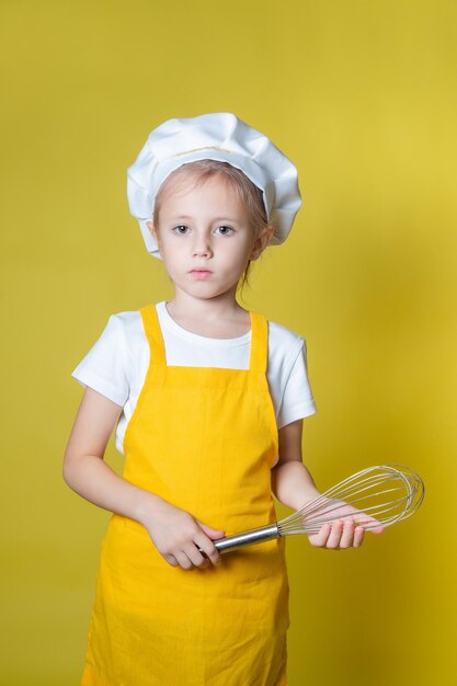 Foto piccola ragazza caucasica che gioca a fare lo chef, ragazza in grembiule e cappello da chef che tiene una frusta per montare la panna su sfondo giallo