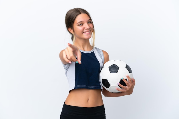 Маленькая кавказская девочка играет в футбол на белом фоне, указывая вперед со счастливым выражением лица