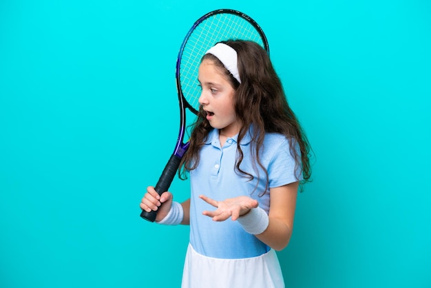 Little caucasian girl playing tennis isolated on blue background with surprise facial expression