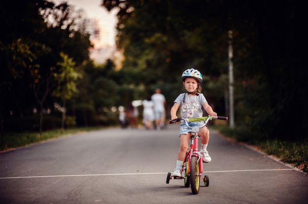 小さな白人の女の子が夏の公園で子供用ヘルメットスニーカーで自転車に乗る