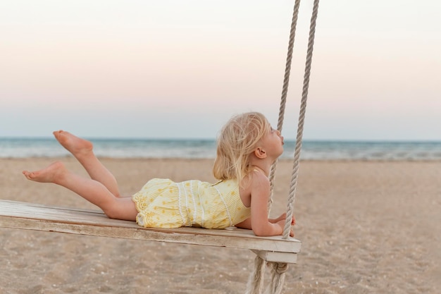 小さな白人の女の子は海の背景にロープスイングに乗る日没時にビーチでかわいい金髪の幼児