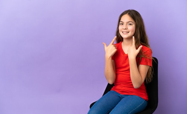 Little caucasian girl sitting on a chair isolated on purple background giving a thumbs up gesture