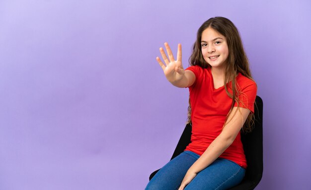 Little caucasian girl sitting on a chair isolated on purple background happy and counting four with fingers