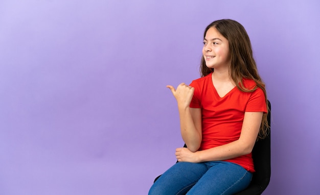 Little caucasian girl sitting on a chair isolated on purple background pointing to the side to present a product