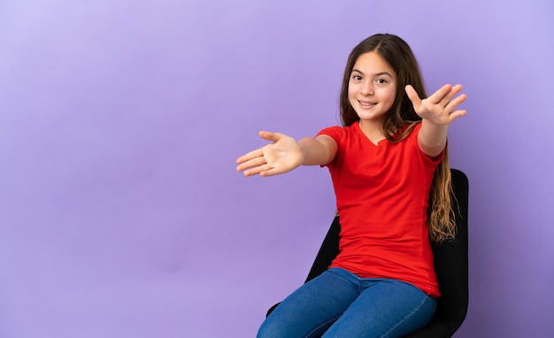 Little caucasian girl sitting on a chair isolated on purple background presenting and inviting to come with hand