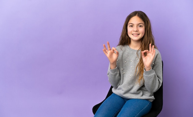 Little caucasian girl sitting on a chair isolated on purple background showing ok sign with two hands