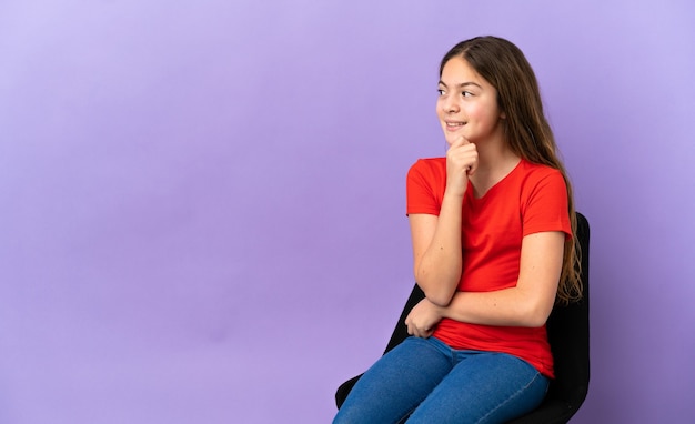 Little caucasian girl sitting on a chair isolated on purple background thinking an idea while looking up