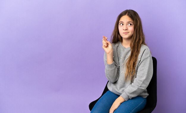 Little caucasian girl sitting on a chair isolated on purple background with fingers crossing and wishing the best